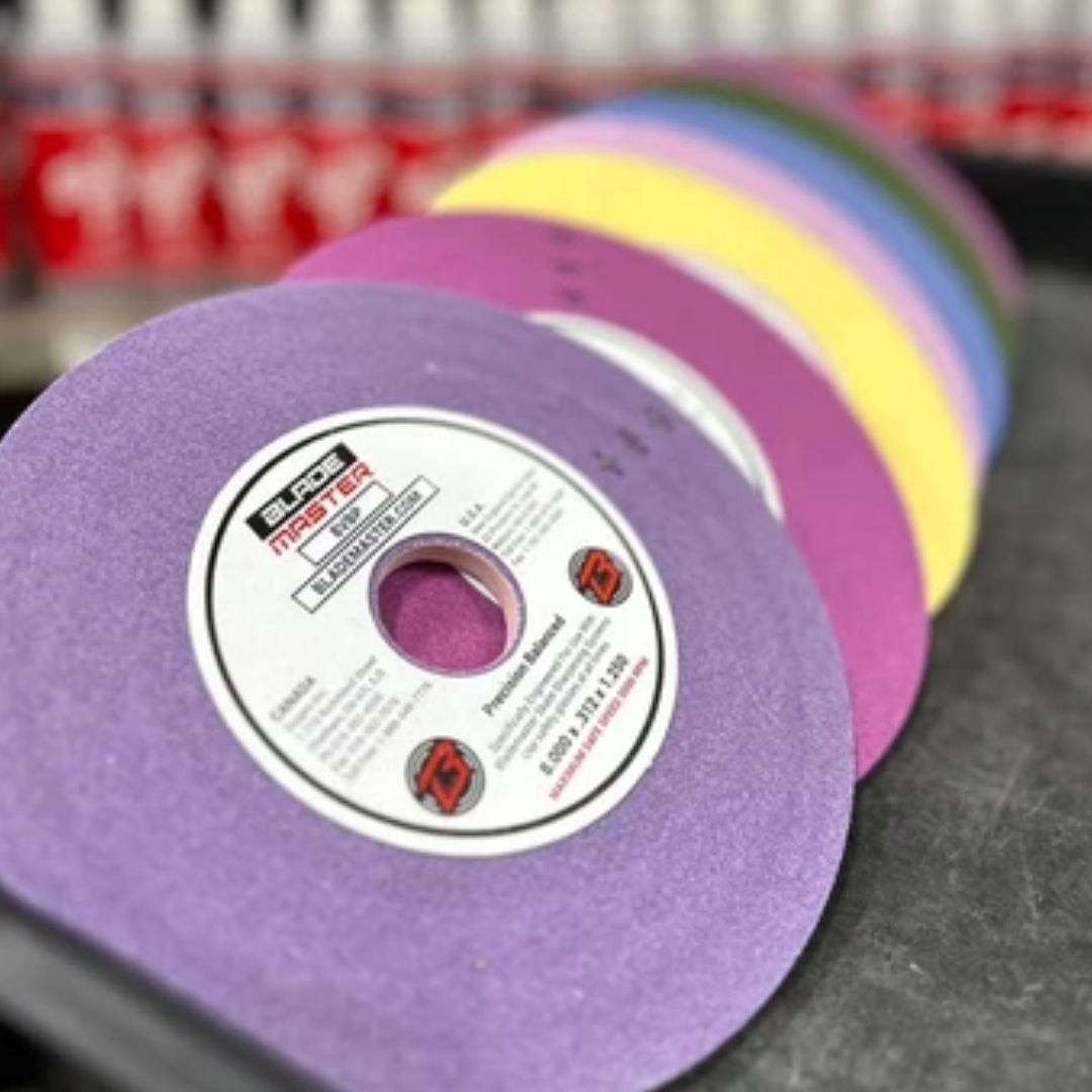 A close-up of a purple grinding wheel with a white label, shown in front of a row of colorful grinding wheels in pink, yellow, green, blue, and white on a dark work surface.